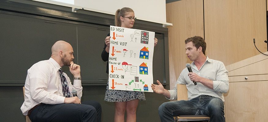 A woman holds up a research poster as two men look on