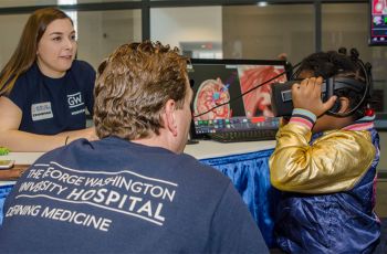 Two GW medical students sit with a child wearing a virtual reality headset