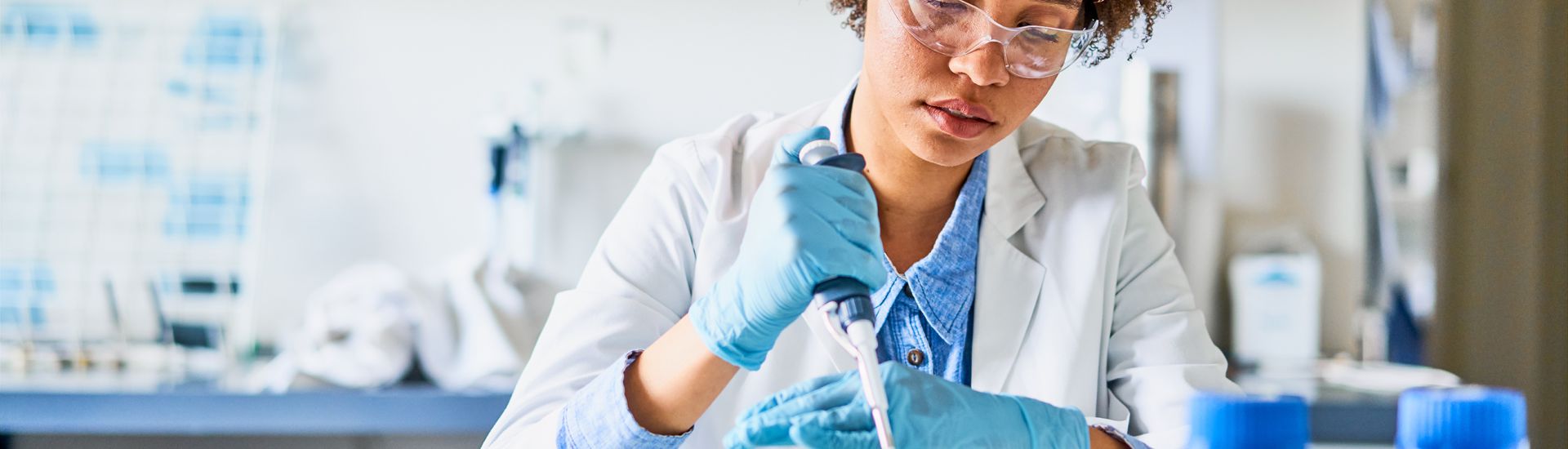 Woman in lab coat, safety goggles, and gloves, using a pipette in a lab.