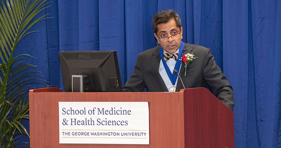 Dr. Raj Rao speaking from a podium wearing a medal and a boutonnière
