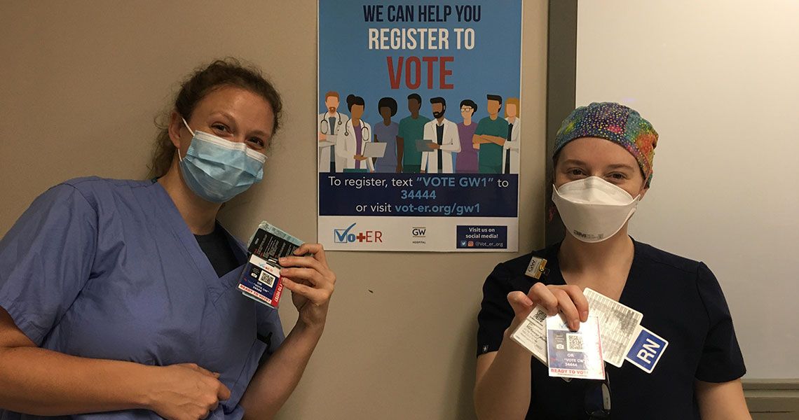 Two medical personnel standing next to a poster that reads 'We can help you register to vote'