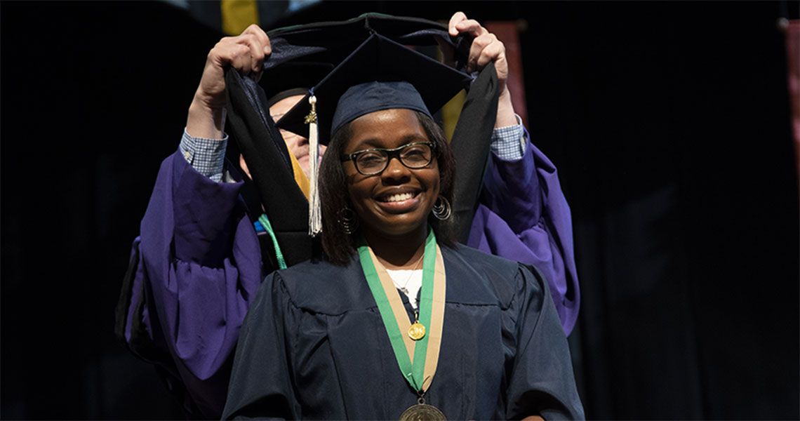 A GW Health Sciences student wearing graduation regalia receives a sash from a faculty member