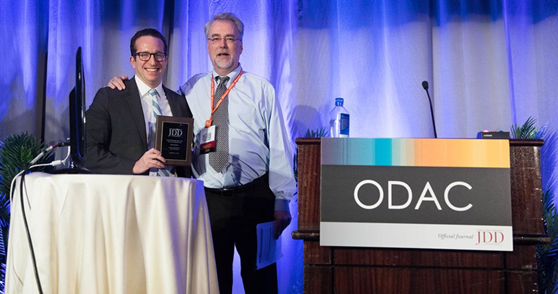 Dr. Adam Friedman holding an award on stage with a man next to a podium that reads 'ODAC'