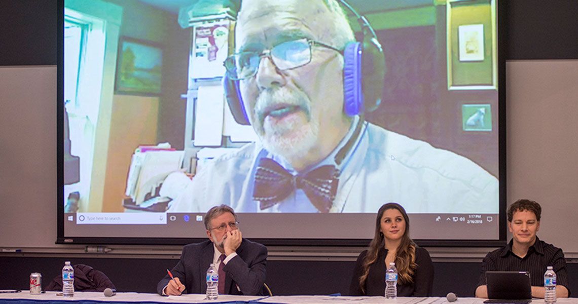 Three GW professors sitting at a table in front of a screen displaying a video call