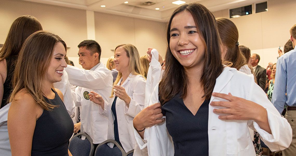 A student putting on a white coat and smiling in a large room of fellow students
