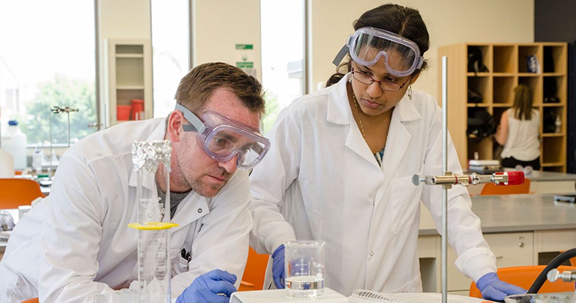 Students in white coats at a table weighing a beaker of liquid
