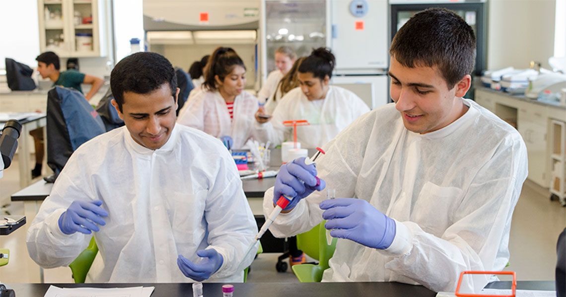 Two high school students wearing white coats and gloves participating in a medical laboratory sciences summer immersion program