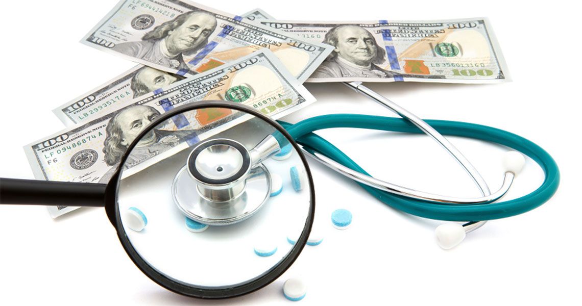 Several one hundred dollar bills next to a stethoscope, magnifying glass and scattered pills