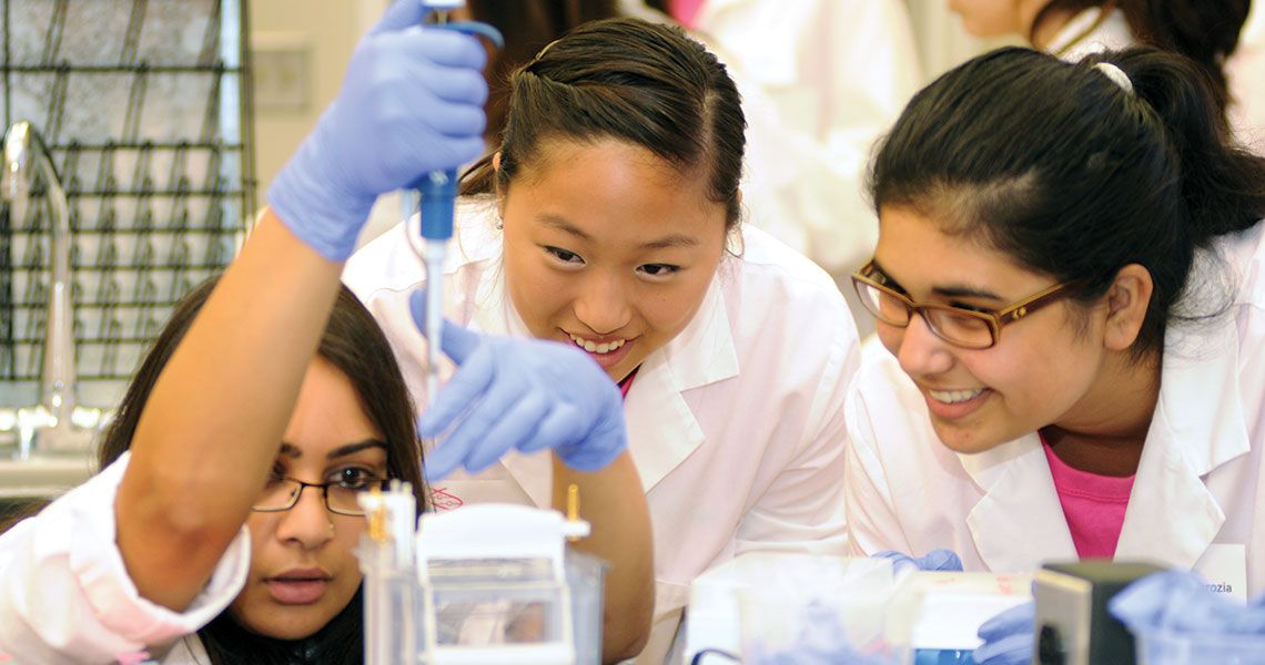 Two females in white coats observing another female in a white coat using a pipette in a laboratory