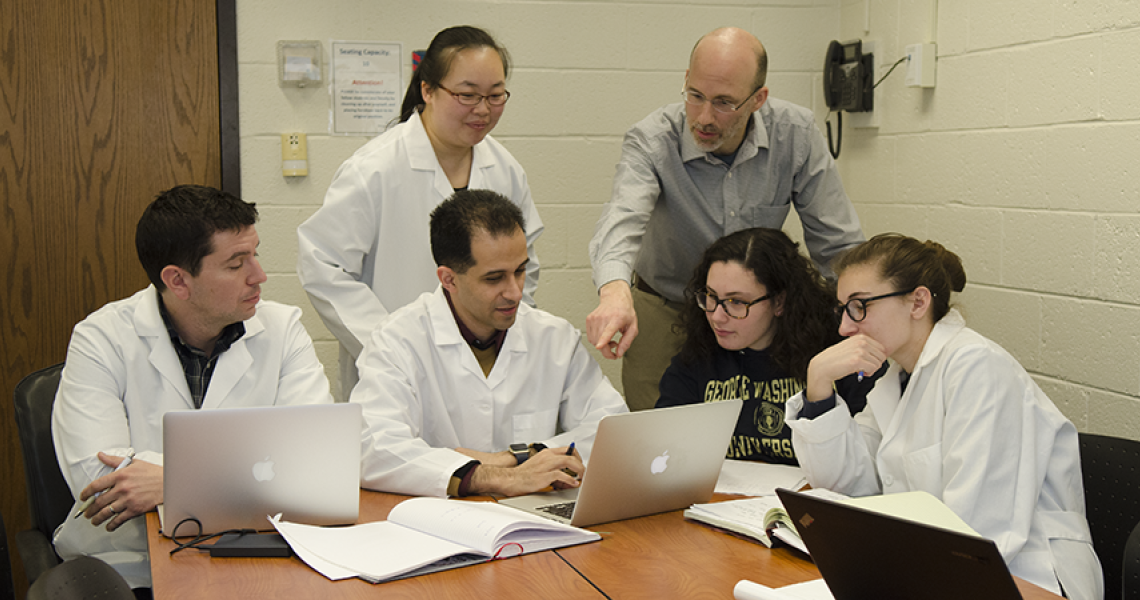 Six people look at a laptop while in a conference room