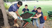 Four people move a wheelbarrow filled with dirt