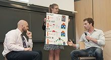 A woman holds up a research poster as two men look on