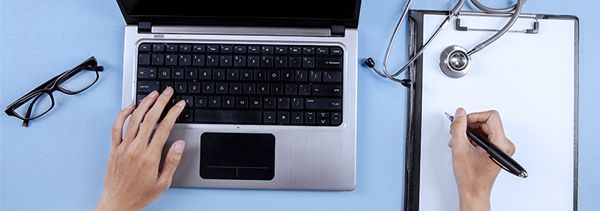 overhead image of laptop with a clipboard, stethoscope, and glasses on the desk next to it