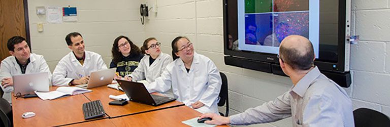 SMHS students in white coats sitting around a table looking at a TV screen