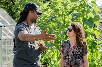 A man gestures in conversation with a woman in front of a greenhouse and garden