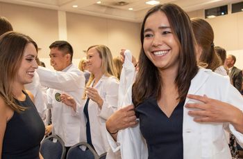 A student putting on a white coat and smiling in a large room of fellow students