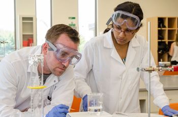 Students in white coats at a table weighing a beaker of liquid